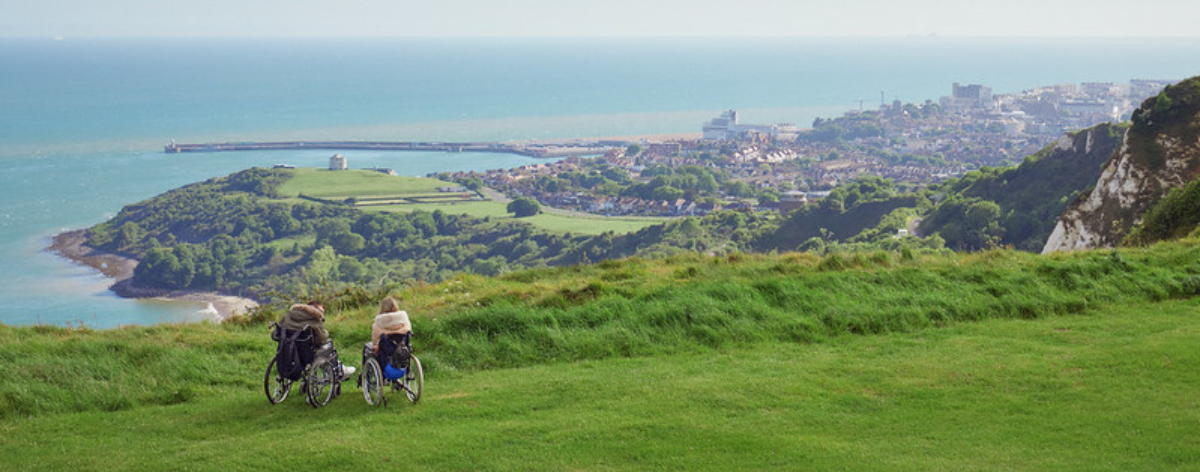 Two people in wheelchairs on a cliff overlooking a coastal view