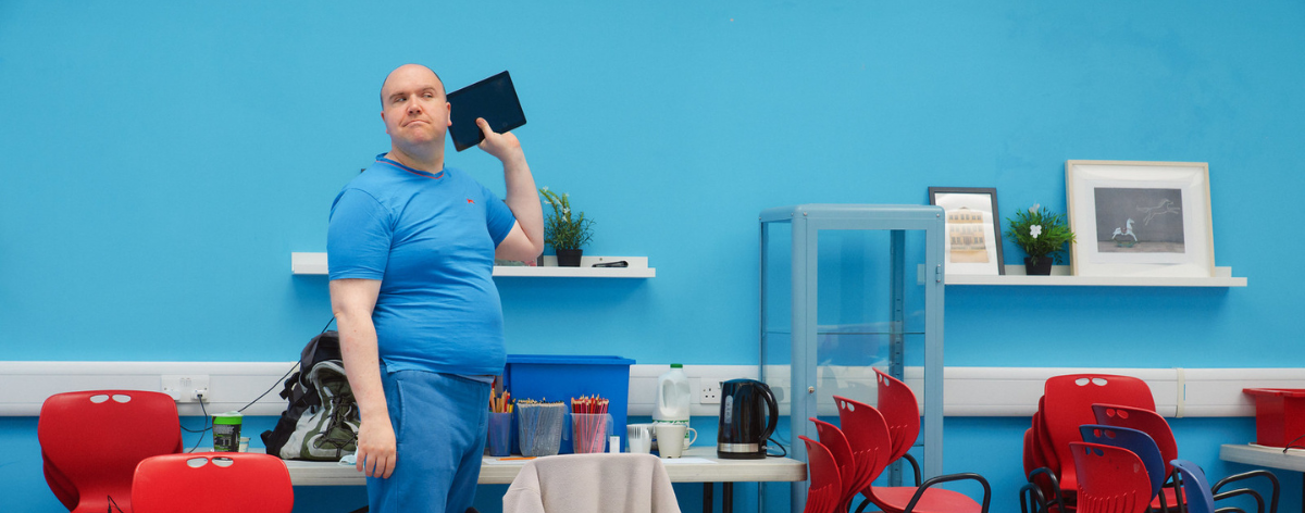 Man in blue T-shirt holding a tablet up to his ear next to a table with tea and coffee making facilities